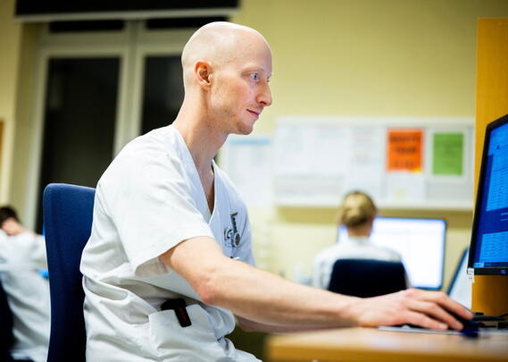 En sykepleier sitter og jobber på en pc i rom med flere sykepleiere.